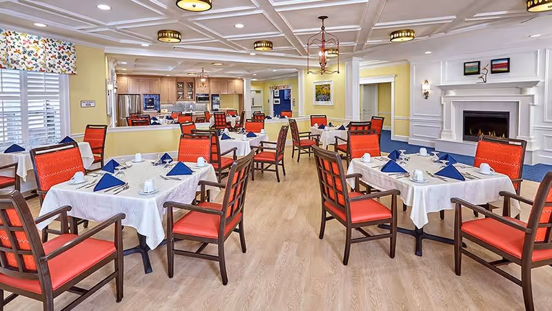 A bright and spacious dining room with multiple tables covered in white tablecloths, each set with blue folded napkins and white cups. The room features wooden chairs with red cushions, a fireplace on the right wall, and a kitchen area visible in the background. The walls are painted yellow and white, and the floor is light wood.