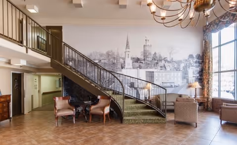 Interior view of a senior living facility lobby with a curved staircase covered in green carpet. There are two upholstered chairs and a small round table beneath the staircase. The wall behind the staircase features a large black and white mural of a town skyline. To the right, there are additional seating areas with armchairs and a large window with floral curtains letting in natural light. A chandelier hangs from the ceiling.