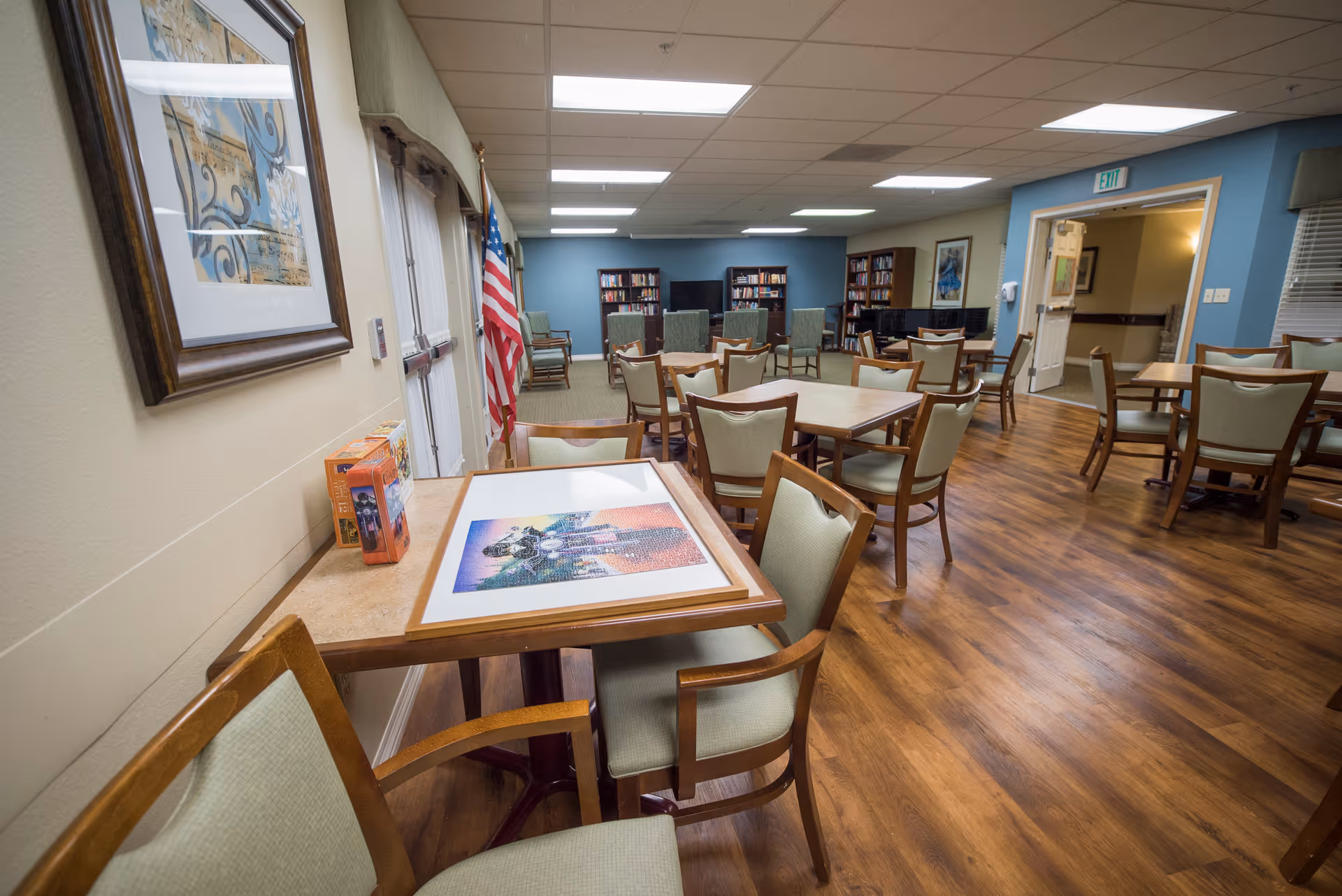 Spacious communal dining/activity room with wooden tables and chairs, bookshelves and an American flag.