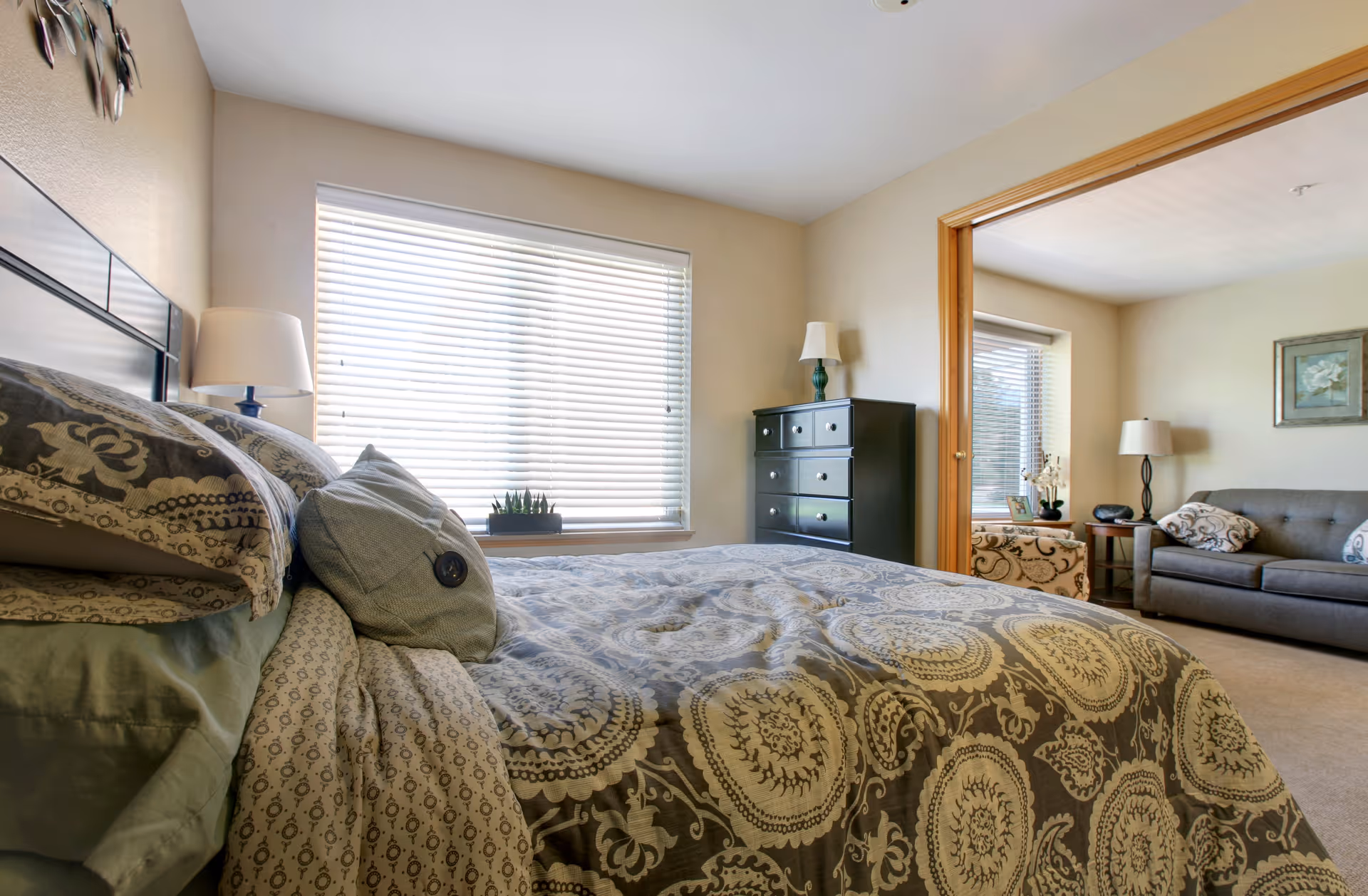 Well-lit bedroom with a patterned bedspread, large window, dresser, and an open doorway into a living area with a sofa.