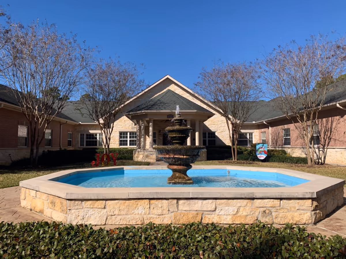 Outdoor courtyard area of a senior living facility with a stone fountain in the center of a blue water pool, surrounded by a stone wall. The building has brick and stone exterior walls with multiple windows and a peaked roof. Leafless trees and some greenery are visible around the courtyard under a clear blue sky.