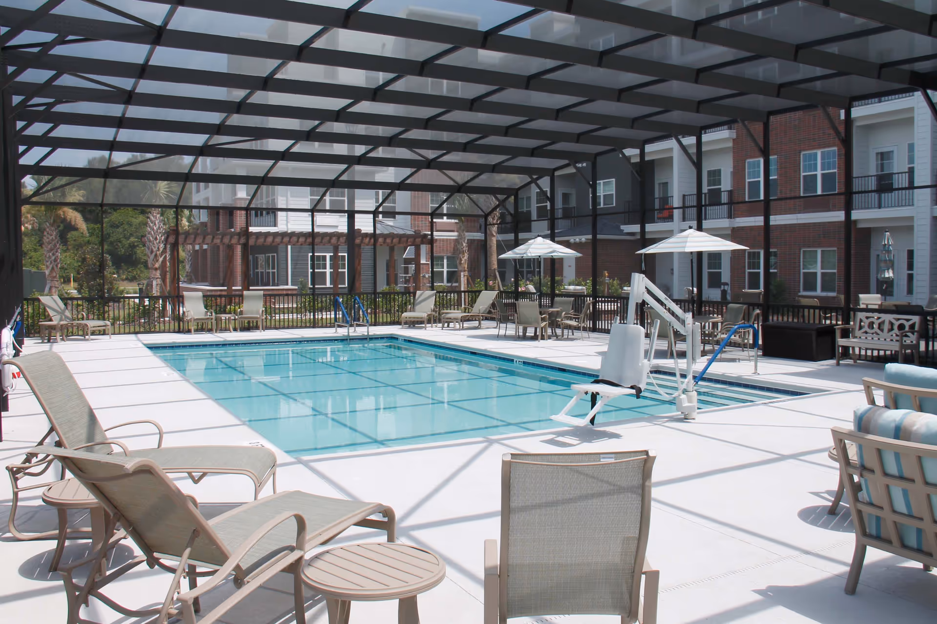 Outdoor swimming pool area enclosed by a screened structure with several lounge chairs, tables with umbrellas, and a pool lift for accessibility. Surrounding the pool are multi-story residential buildings with balconies and windows.