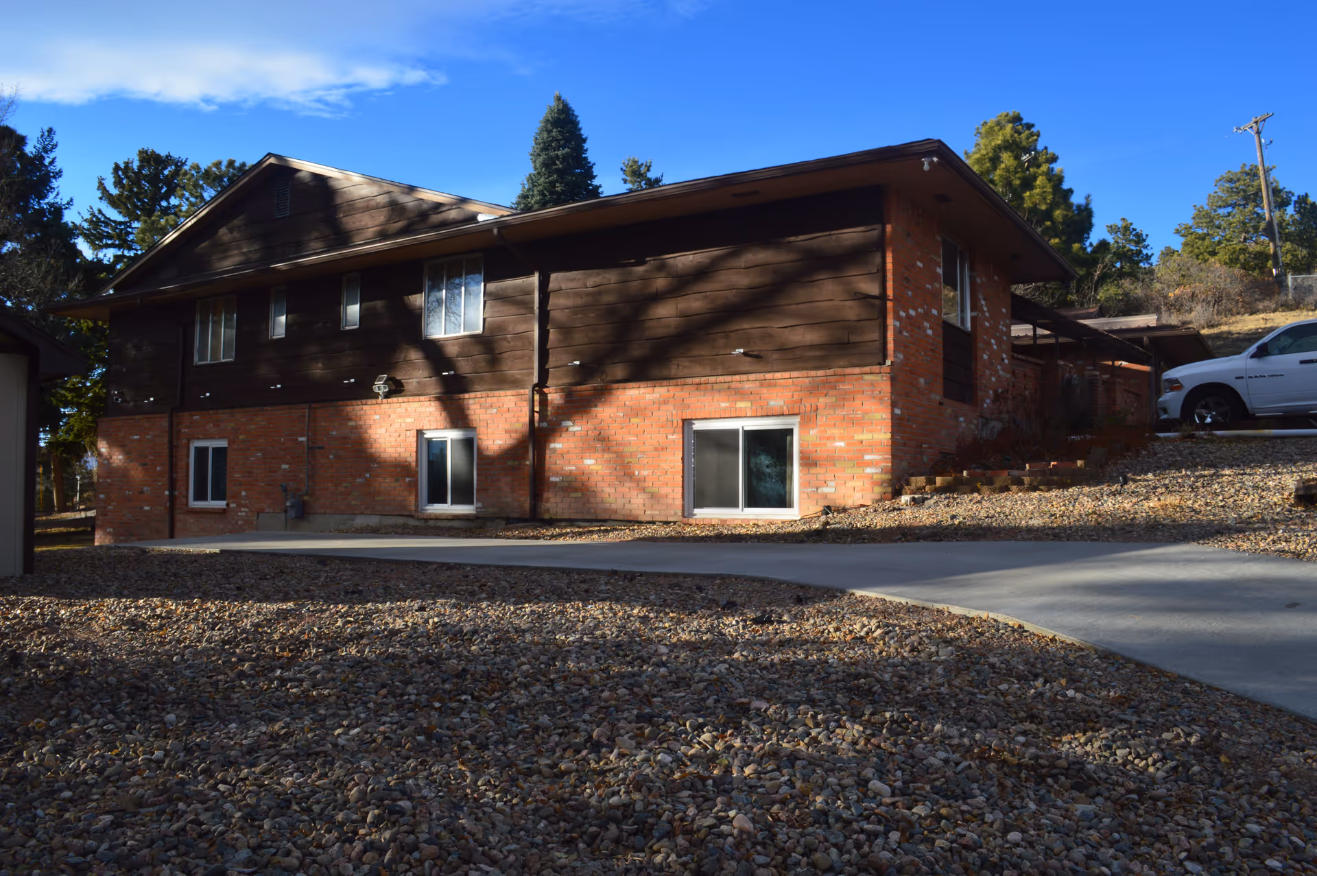 Two-story brick-and-wood building with a gravel front yard, driveway, and a parked white SUV under a clear blue sky.