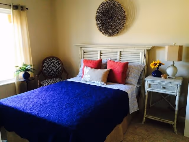 A cozy bedroom with a bed covered in a dark blue quilt, two red pillows and one beige pillow. There is a white wooden headboard with a decorative round wall hanging above it. To the right of the bed is a white nightstand with a lamp and a small vase of sunflowers. To the left of the bed is a window with white curtains and a small round table holding a potted plant next to a patterned armchair.