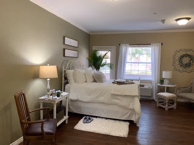 Neatly staged bedroom with a white bed, bedside tables and lamps, chairs, a small rug, and a window with curtains.