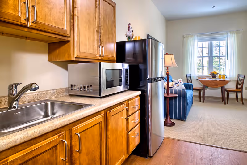 A small kitchen area with wooden cabinets, a stainless steel sink, and a microwave on the countertop. Next to the kitchen is a living space with a blue striped sofa, a floor lamp, and a small wooden dining table with two chairs near a window with white curtains.