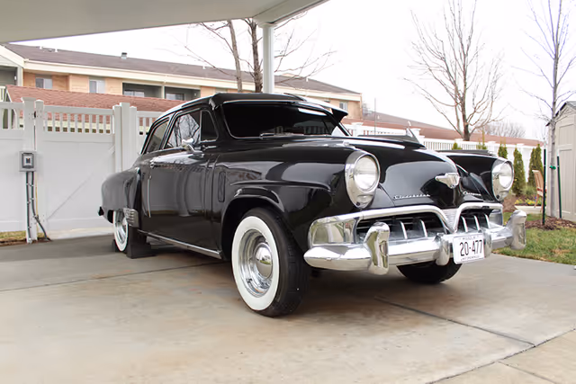 A vintage black car with whitewall tires parked under a covered driveway next to a white fence and some leafless trees in a residential or facility area.