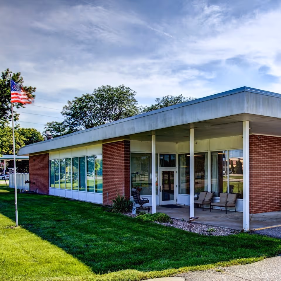Low one-story brick and glass building with a covered entrance, lawn, American flag, and outdoor chairs.