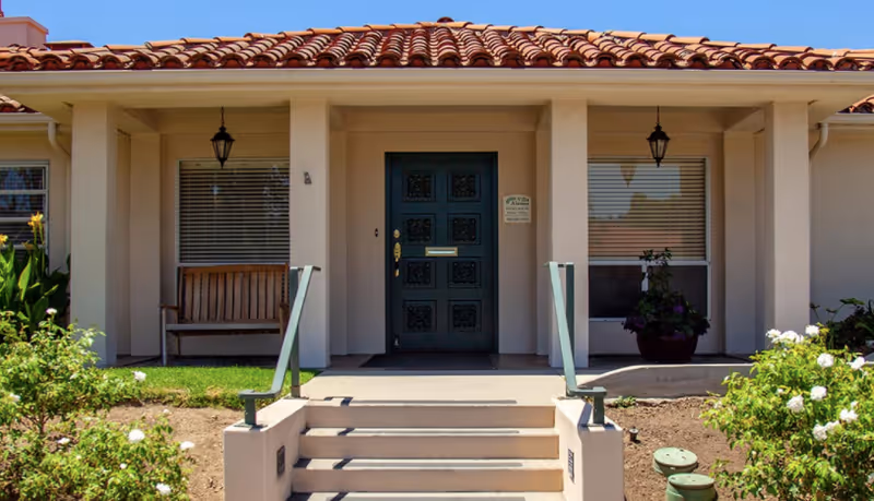 Front entrance of a building with a tiled roof, a dark green door with decorative panels, two hanging lanterns, a wooden bench on the left side, potted plants on the right, and steps leading up to the door surrounded by greenery and white flowers.