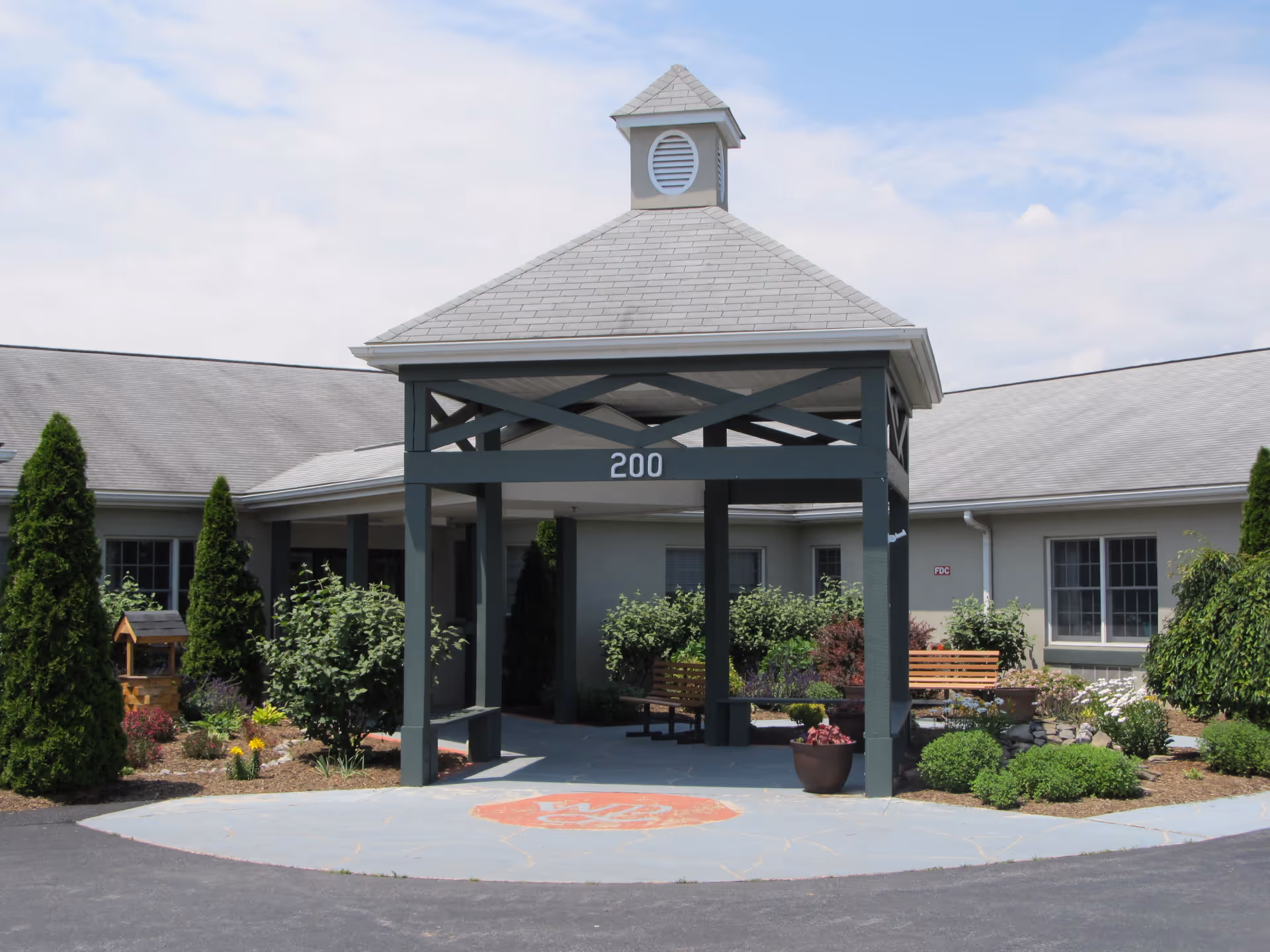 Entrance of Whispering Pines Assisted Living facility featuring a covered porch with the number 200 displayed on the structure. The area is surrounded by well-maintained shrubs, small trees, and flower beds, with benches placed under the porch for seating.