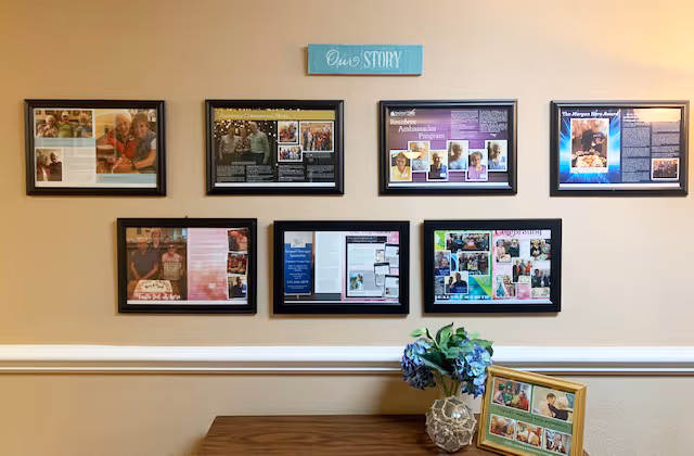 A beige wall with seven framed photos and informational displays arranged in two rows, with a small blue sign above them that reads 'Our Story.' Below the frames is a wooden surface holding a decorative vase with blue flowers and a small framed photo collage.