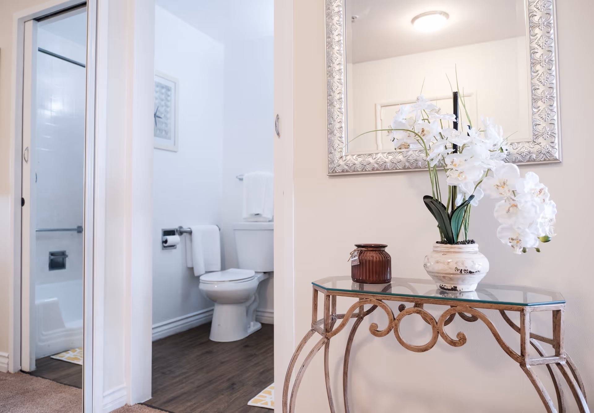 Decorative hallway table with a vase of white orchids and mirror next to an open doorway showing a bathroom with a toilet and shower.