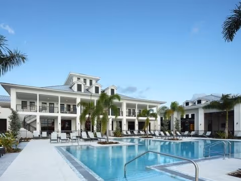 Outdoor view of a senior living facility named Wellington Bay featuring a large swimming pool surrounded by lounge chairs and palm trees, with a two-story building in the background under a clear blue sky.