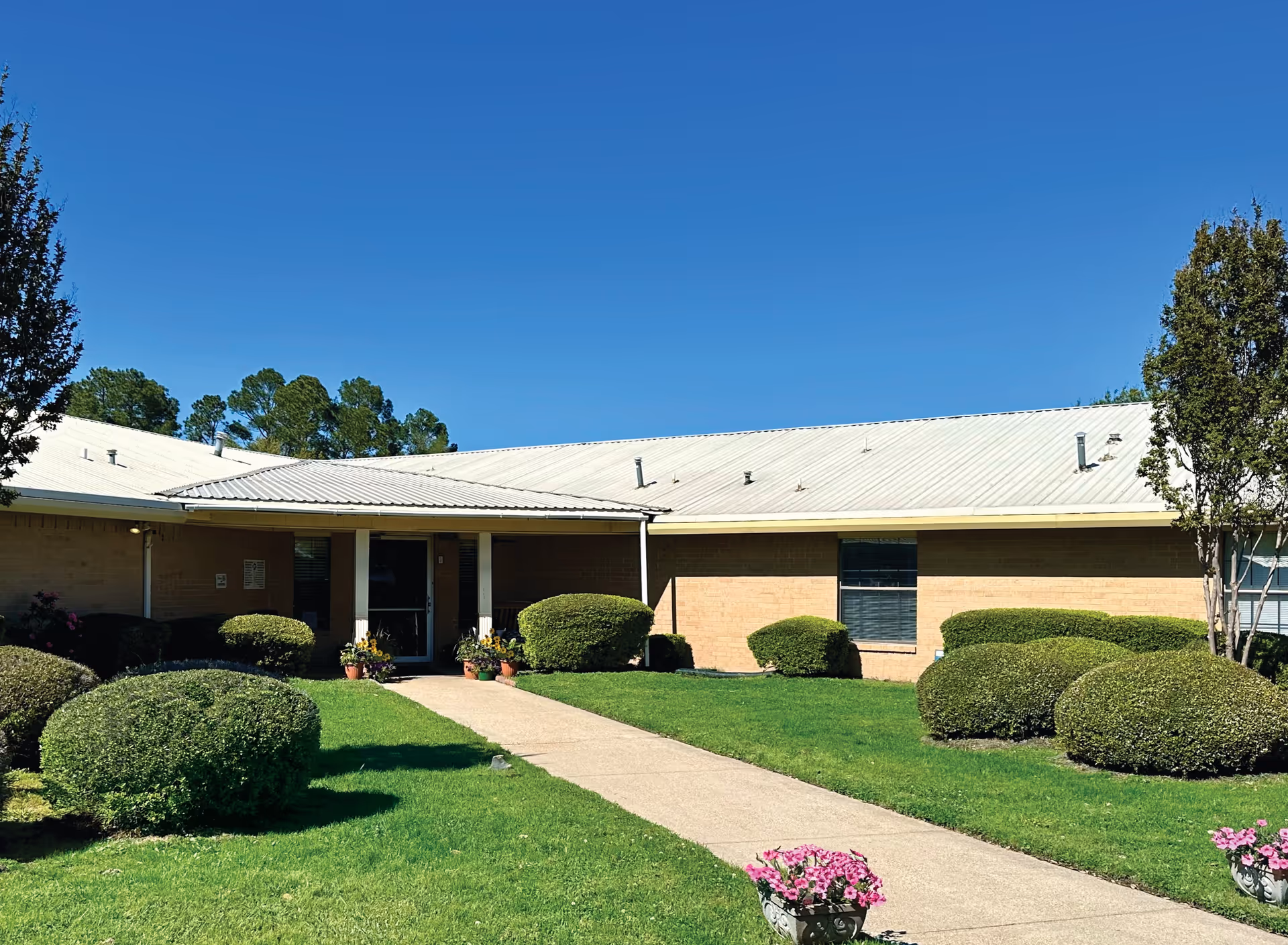 Front entrance of a single-story brick senior living facility with a walkway, manicured shrubs, and flower pots under a clear blue sky.