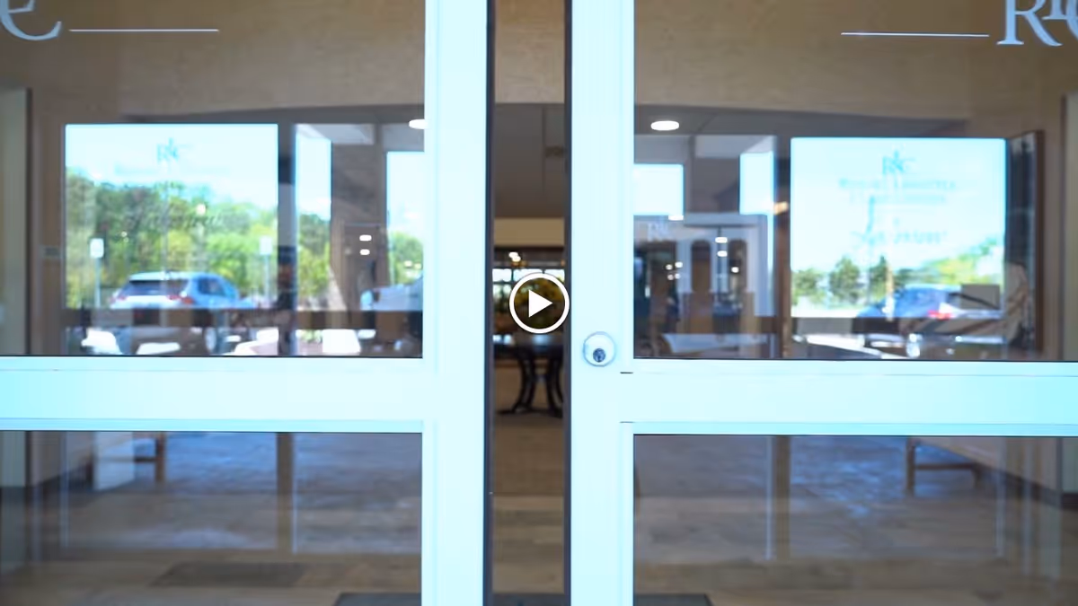 Glass double doors at the entrance of a building with reflections of parked cars and greenery outside. Inside, a hallway with a table and chairs is visible.
