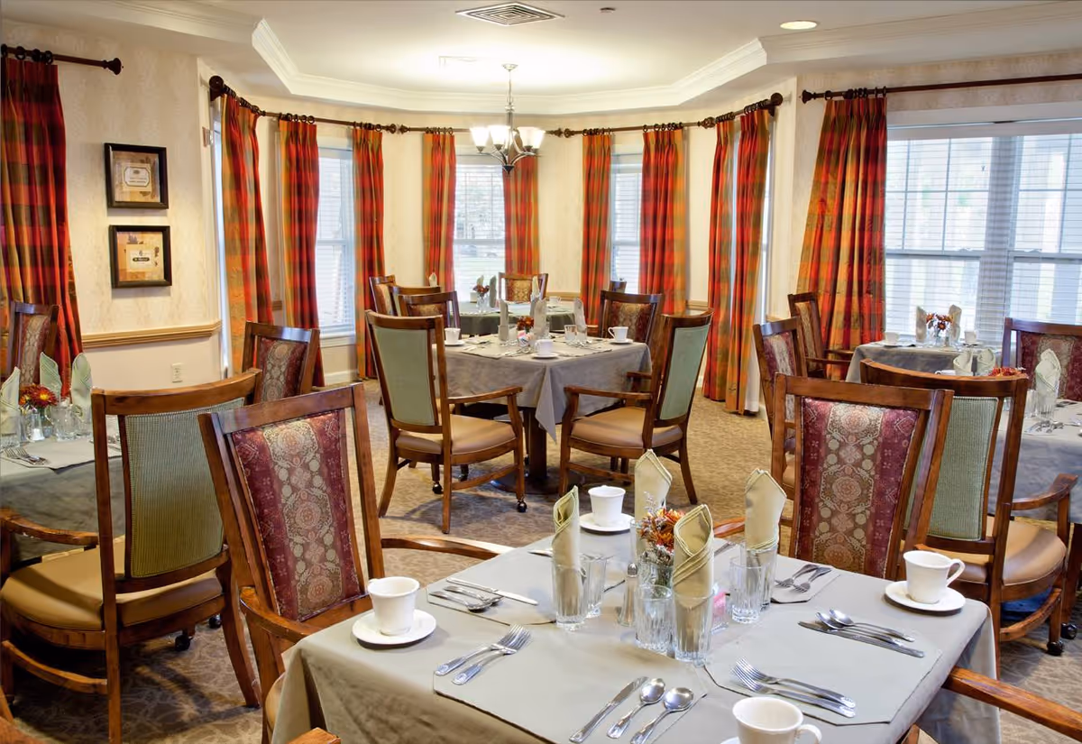 Dining room with several tables set for a meal, wooden chairs, and tall windows framed by red and orange plaid curtains.