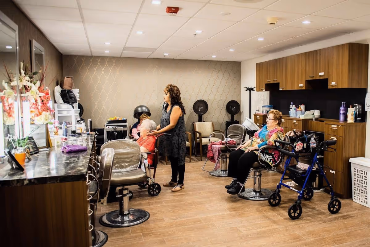 A hair salon inside a senior living facility with a stylist attending to an elderly woman seated in a salon chair. Another elderly woman is seated nearby with a walker beside her. The room has wooden flooring, a counter with flowers and hair products, and cabinets along the wall.