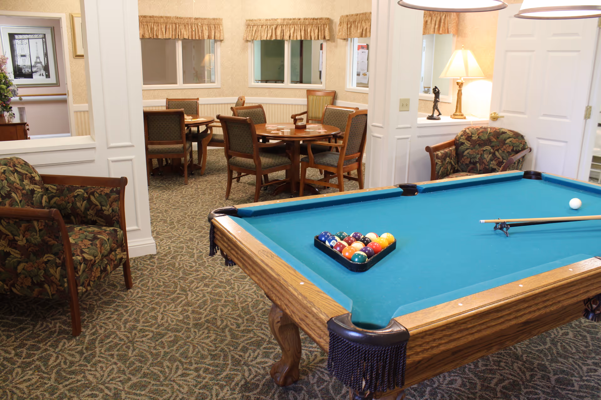 A senior living facility common room with a pool table in the foreground and seating and card tables in the background.