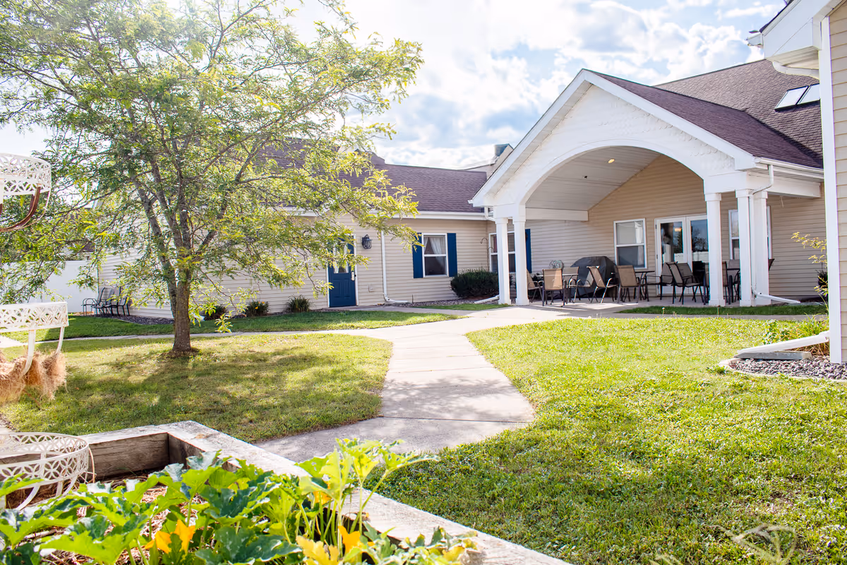 Outdoor view of a senior living facility courtyard with a paved walkway, green grass, a tree, garden plants, and a covered patio area with chairs and tables attached to a beige building with blue doors and windows.