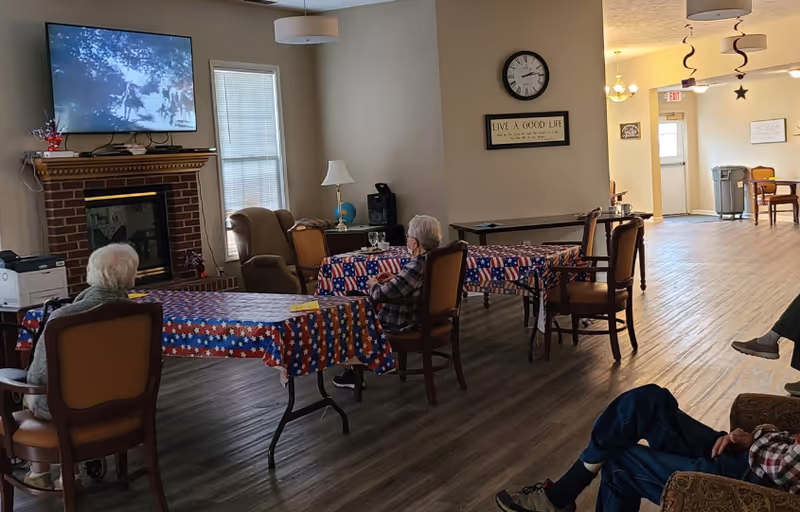 Several elderly residents sit at tables with star-spangled tablecloths in a communal lounge watching a television above a fireplace.