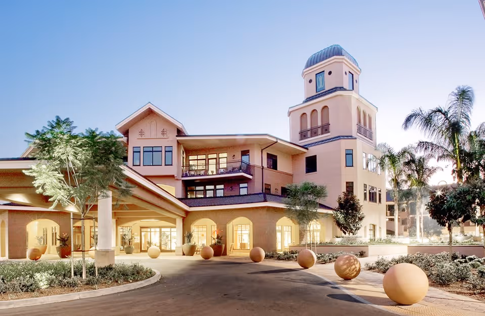 Exterior view of Walnut Village Orange County Retirement Community building at dusk, featuring a multi-story structure with a tower, large windows, a covered entrance, palm trees, and landscaped grounds with spherical decorative elements.