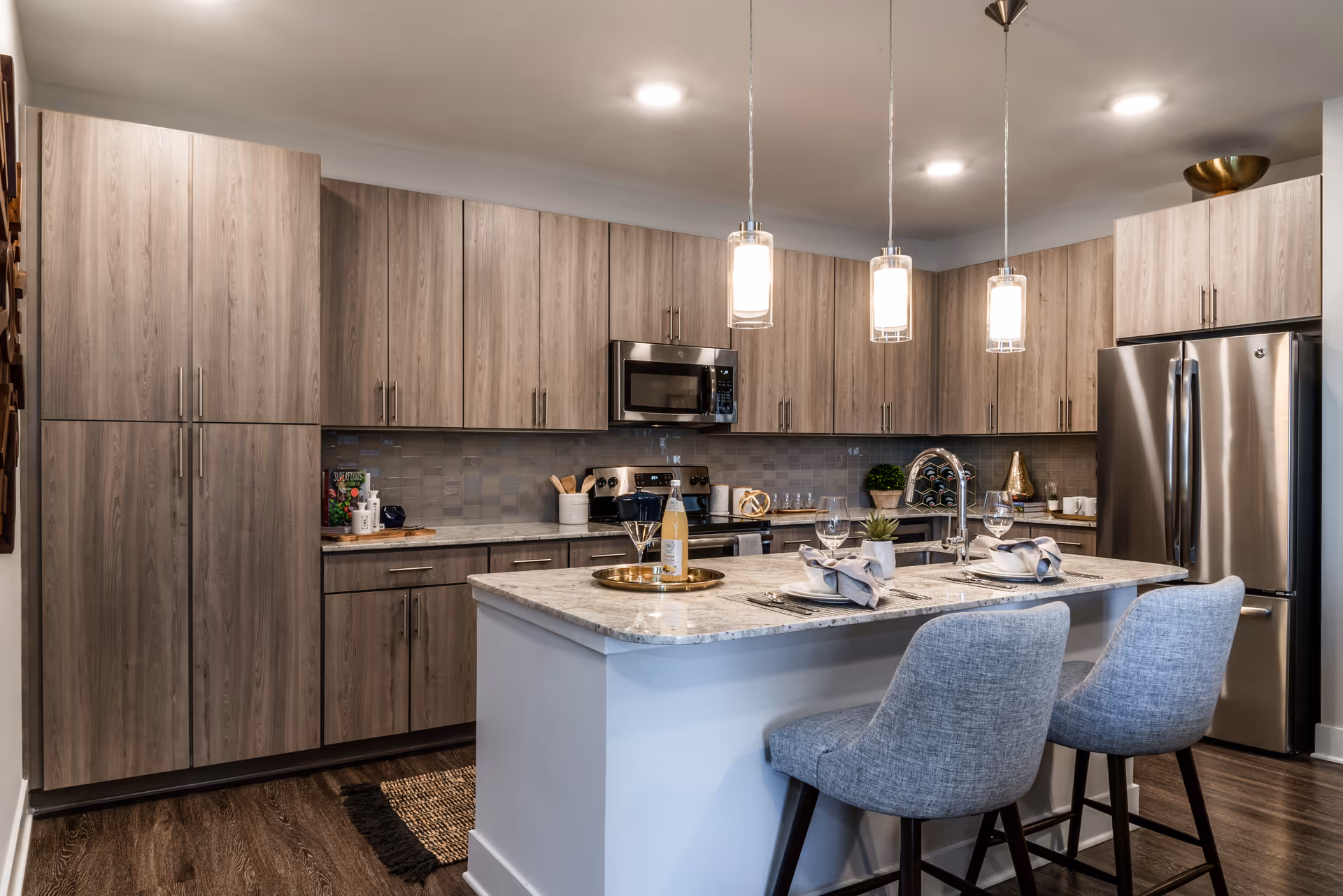 Modern kitchen with a marble island, two upholstered bar stools, stainless steel appliances and wood grain cabinets.