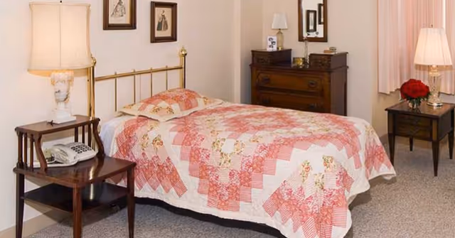 Cozy bedroom with a single brass bed covered by a pink floral quilt, bedside tables with lamps, a wooden dresser, and wall art.