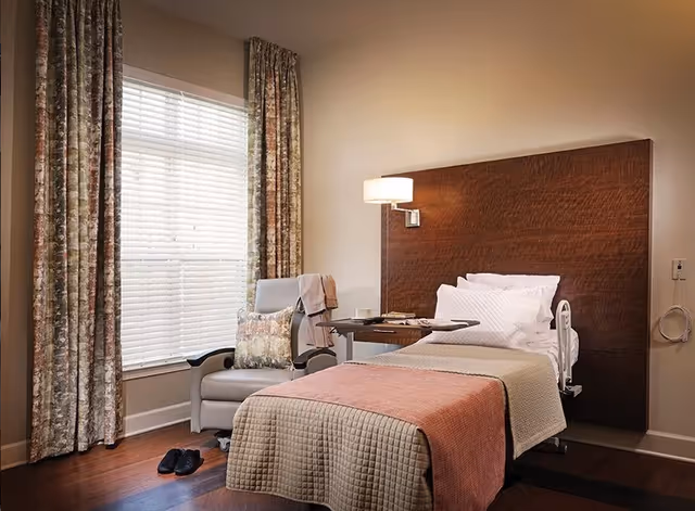 A neatly made single hospital-style bed with a wooden headboard, bedside table and lamp, and an armchair by a window with patterned curtains in a cozy senior living bedroom.