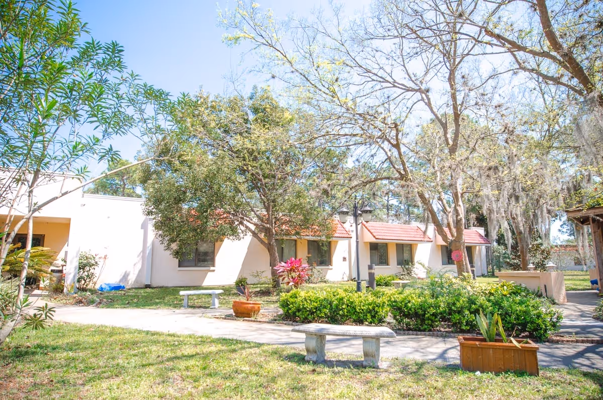 Sunlit courtyard with benches, potted plants, and trees in front of single-story nursing facility buildings with red-tiled roofs.