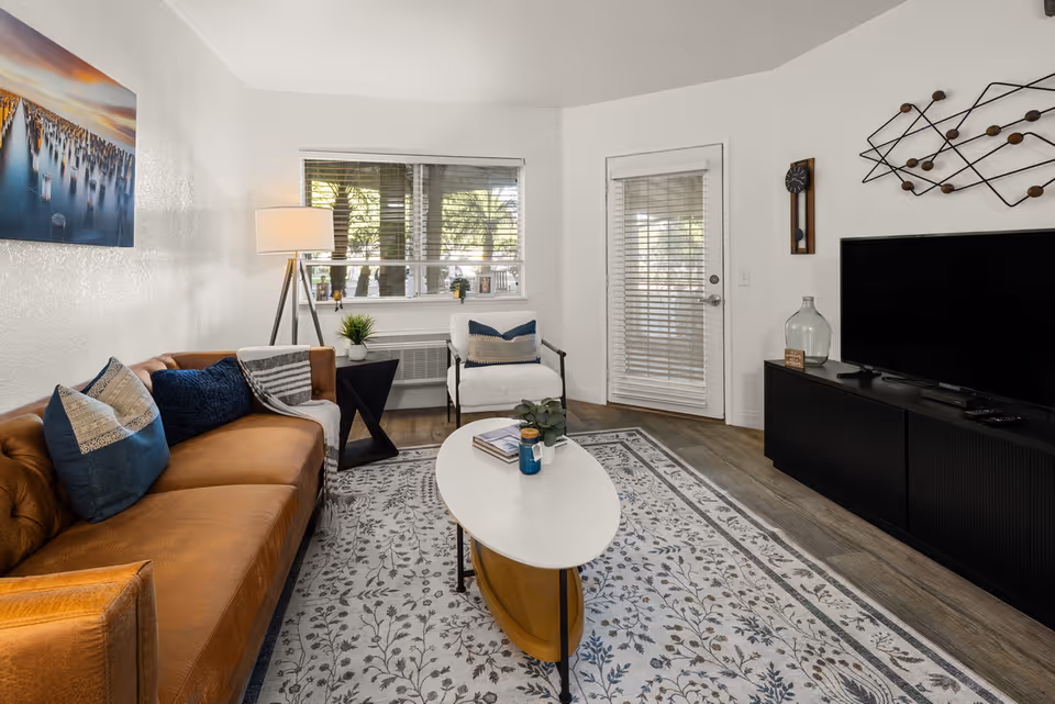 A cozy living room with a brown leather sofa adorned with blue and beige pillows, a white armchair, a white oval coffee table with books and a small plant, a patterned area rug, a floor lamp, a large flat-screen TV on a black media console, and wall decorations including a clock and abstract metal art. A window with blinds and a glass door with blinds allow natural light into the room.