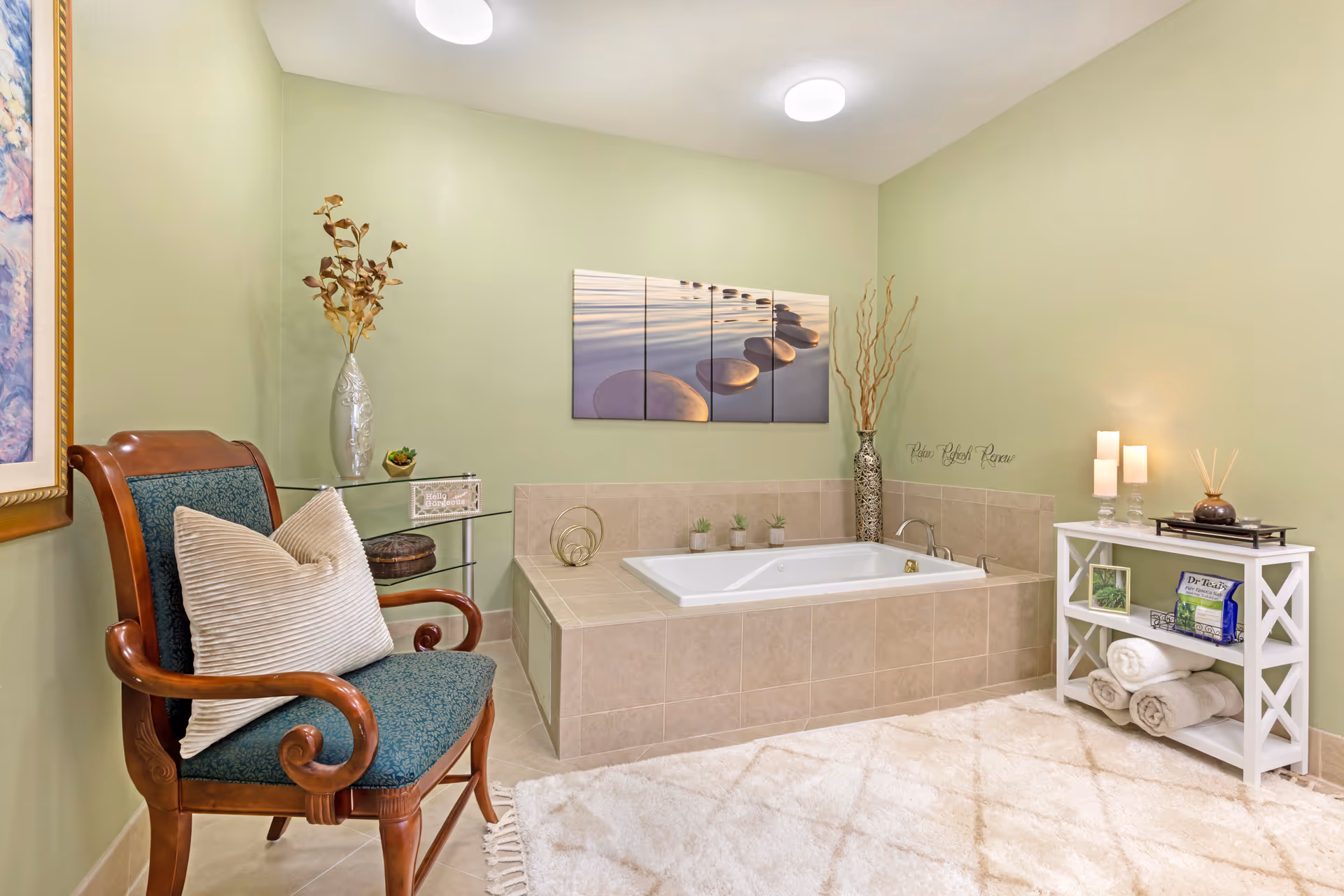 A serene bathroom featuring a large built-in bathtub with beige tiles, decorated with small potted plants and a tall vase with dried branches. A wooden chair with green upholstery and a beige pillow sits next to a glass shelf holding decorative items. On the right, a white shelving unit holds rolled towels, candles, and a box of tea. The walls are painted light green, and a multi-panel artwork of smooth stones on water hangs above the bathtub. A plush white rug covers the floor.