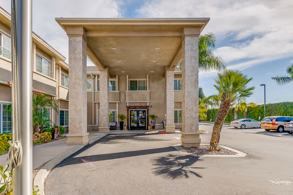 The covered front entrance/porte-cochere of a residential building with large columns, palm trees, and parked cars in the driveway.