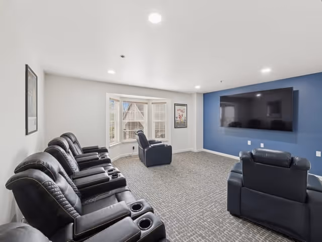 A modern lounge room with multiple black leather recliner chairs arranged facing a large flat-screen TV mounted on a blue accent wall. The room has a carpeted floor, white walls, recessed ceiling lights, and a bay window with white blinds letting in natural light. Two framed pictures hang on the walls.
