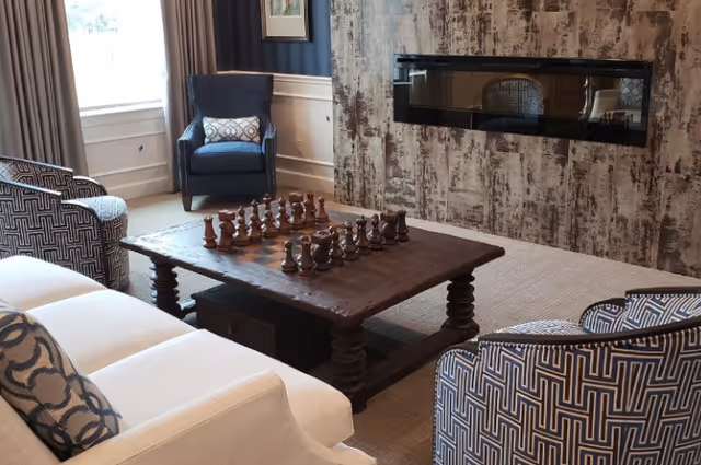 A cozy living room with a wooden coffee table featuring a chess set, surrounded by a white sofa and patterned armchairs. A blue armchair with a decorative pillow is placed near a window with curtains. The room has a textured wall with a modern built-in fireplace.