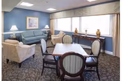 A cozy sitting area in a retirement community featuring a blue sofa, two patterned armchairs, a wooden table with four chairs, two table lamps on side tables, decorative vases on a console table by large windows, and framed artwork on the wall.