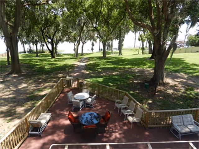 Wooden deck patio with tables and chairs overlooking a grassy yard dotted with large shade trees.