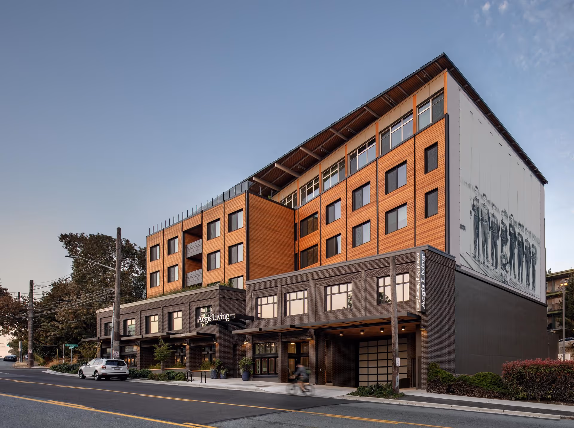 Exterior view of a modern multi-story building with a combination of dark brick and wooden paneling. The building has large windows and a sign that reads 'Aegis Living'. A person on a bicycle is passing by on the street in front of the building, and a white car is parked on the side of the road. The sky is clear with a few clouds.