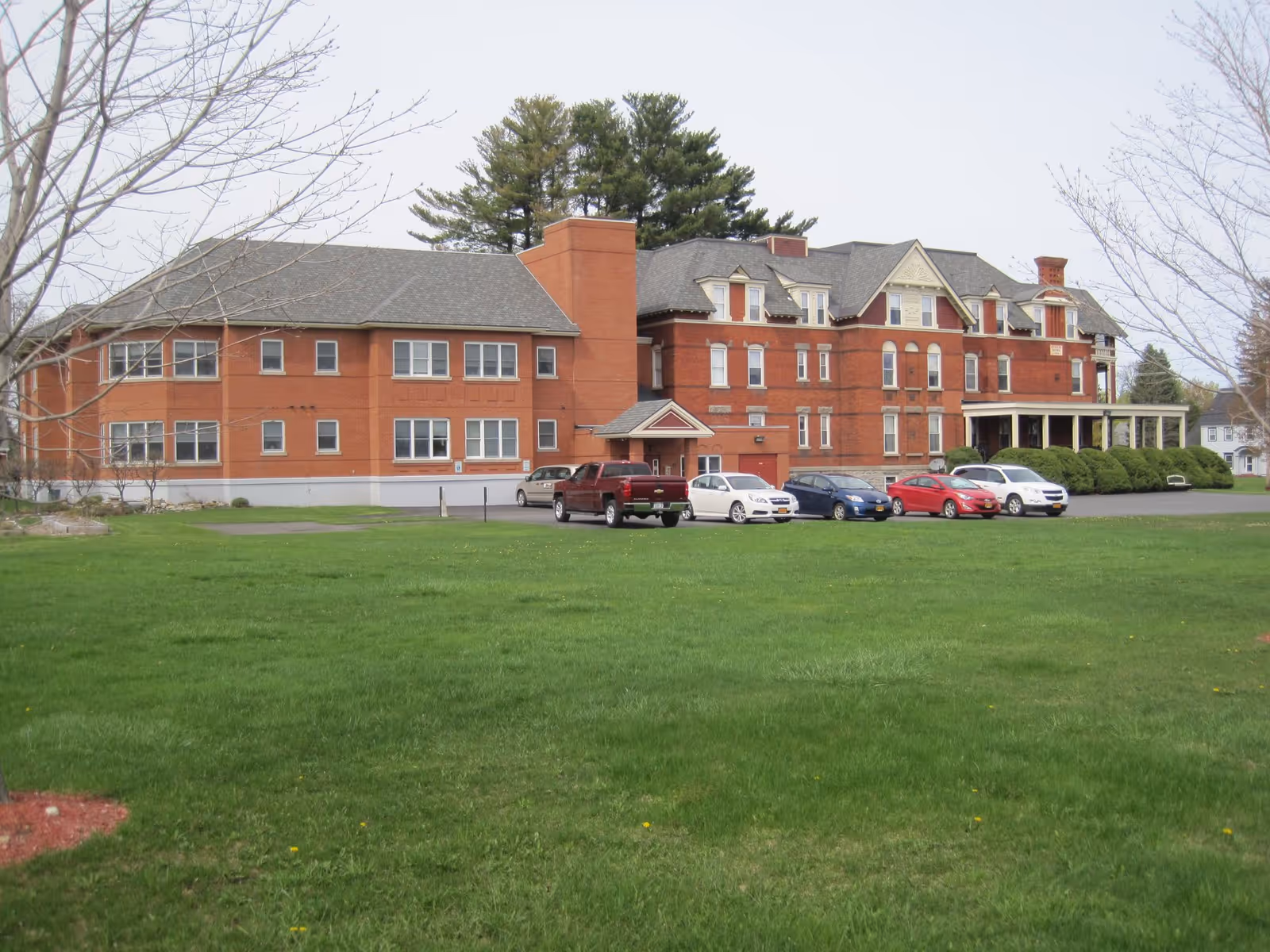 Large red-brick multi-story building with a grassy lawn and parked cars in front.