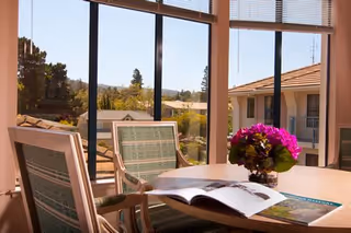 Sunlit dining table with chairs, an open magazine and a vase of purple flowers by large windows overlooking the courtyard and neighboring building.