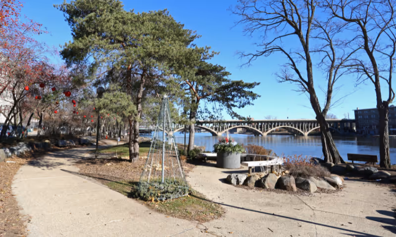 Riverside park walkway with trees, benches, planters and a bridge crossing the river under a clear blue sky.