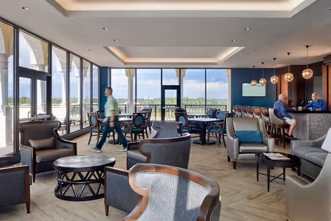 A spacious and well-lit lounge area with large floor-to-ceiling windows offering a view of the outdoors. The room features multiple comfortable armchairs and round tables arranged for socializing. In the background, two people are seated at a bar counter with hanging pendant lights above. A person is walking through the space, and the overall atmosphere is modern and inviting.