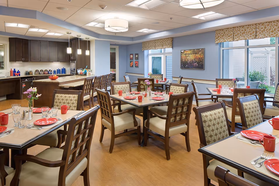 A bright and spacious dining room in a senior living facility with several tables set with red plates, red mugs, silverware, and glasses. The room features wooden chairs with patterned cushions, a kitchen area with dark cabinets and pendant lights, large windows with patterned valances, and decorative artwork on the walls.
