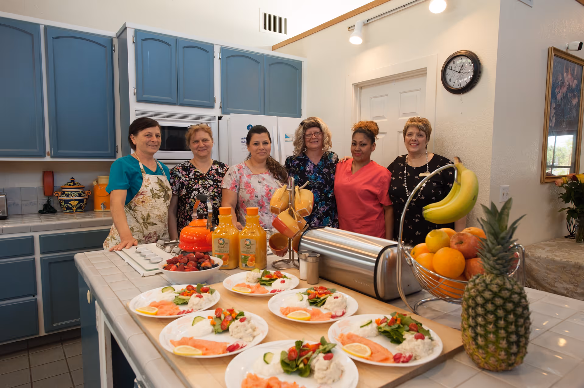 Six women standing behind a kitchen counter with plates of food, bottles of orange juice, a bowl of strawberries, and a fruit basket containing bananas, oranges, apples, and a pineapple. The kitchen has blue cabinets and a clock on the wall showing 10:10.