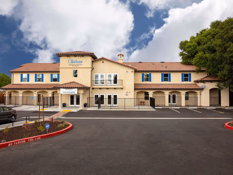 Front exterior view of The Chalet senior living facility, a two-story building with beige stucco walls, red tile roof, blue window shutters, and a parking lot in front under a partly cloudy sky.