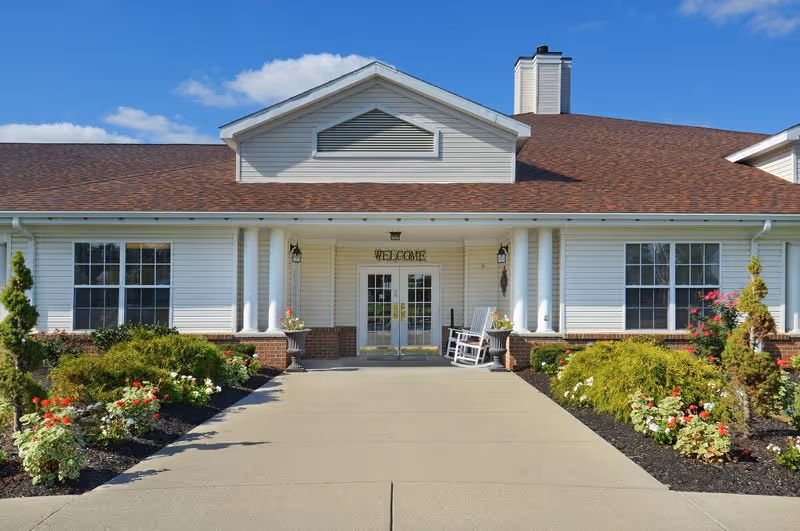 Front exterior view of a single-story senior living facility building with white siding, a brown shingled roof, and a welcoming entrance with double glass doors. There are two white rocking chairs on the porch, flower beds with colorful flowers and shrubs on either side of the concrete walkway leading to the entrance, and a clear blue sky above.