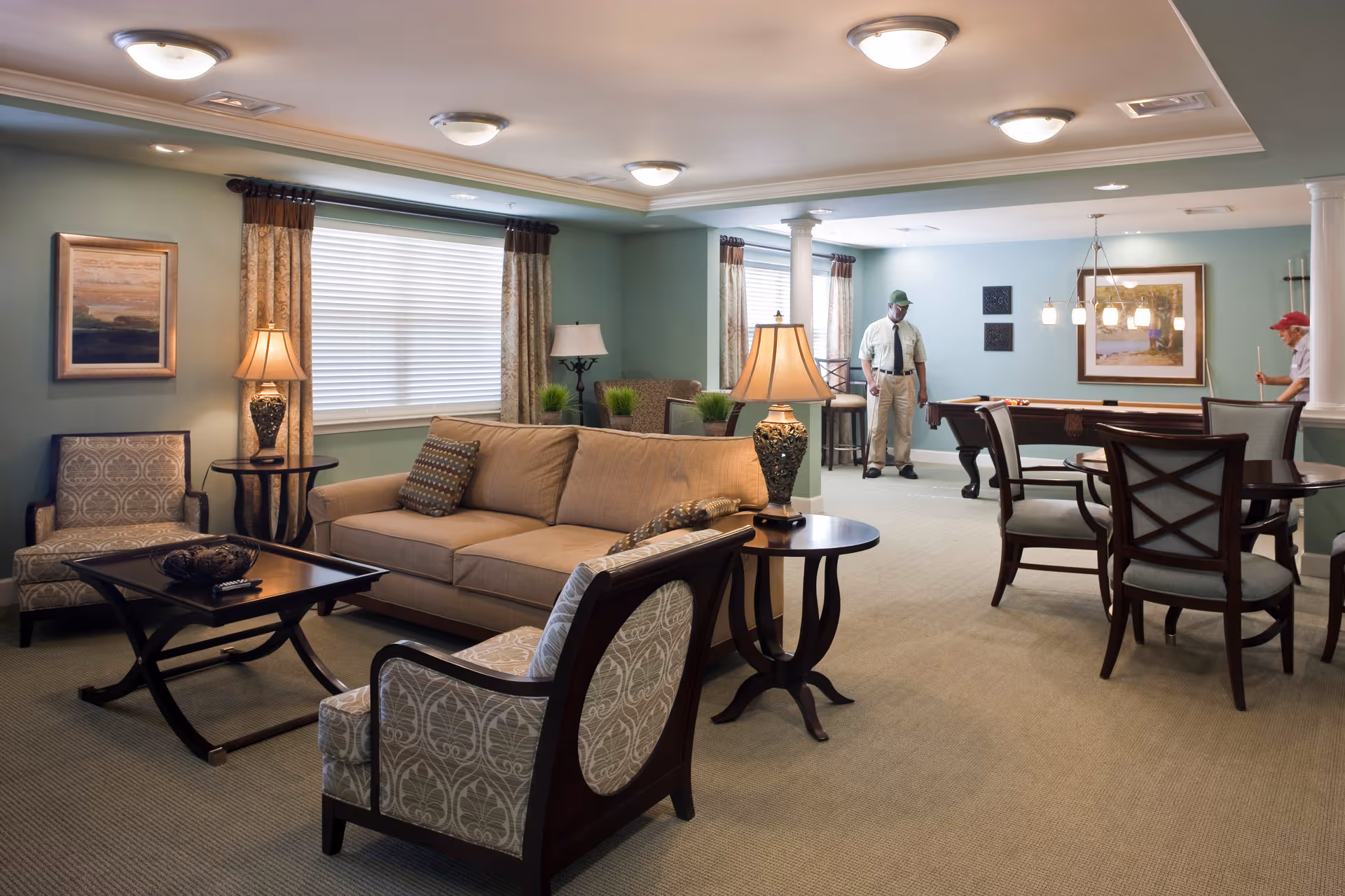 A spacious and well-lit common living area with beige and patterned armchairs, a beige sofa, wooden side tables with lamps, and a coffee table. In the background, two elderly men are playing pool near a pool table under hanging lights. The walls are painted light green with framed artwork and windows with curtains.