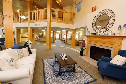 Spacious senior living facility common area with a white sofa and blue armchair around a wooden coffee table on a patterned rug. There is a fireplace with a decorative round mirror above it and candles on the mantel. The room has high ceilings with wooden beams and a balcony railing on the upper level.