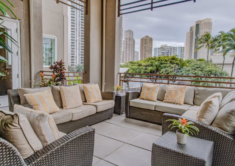 Outdoor patio area with cushioned wicker sofas and chairs arranged around a small wicker table with a potted plant. The patio overlooks a cityscape with tall buildings and lush green trees under a cloudy sky.