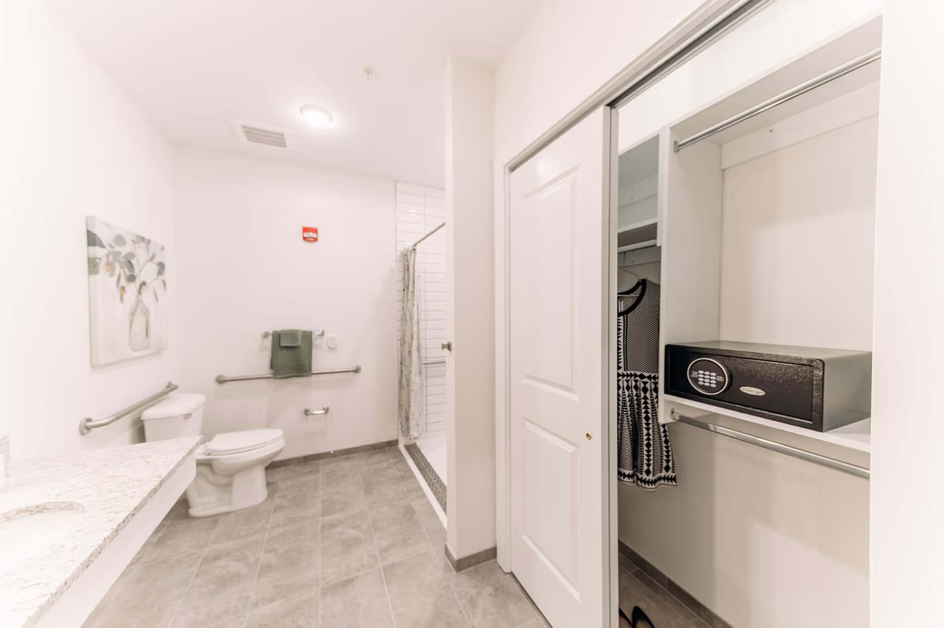 A clean, modern bathroom with a toilet, grab bars, a towel rack with a green towel, a shower with a curtain, and a granite countertop sink. To the right, there is an open closet with a black and white dress hanging and a black safe on a shelf.