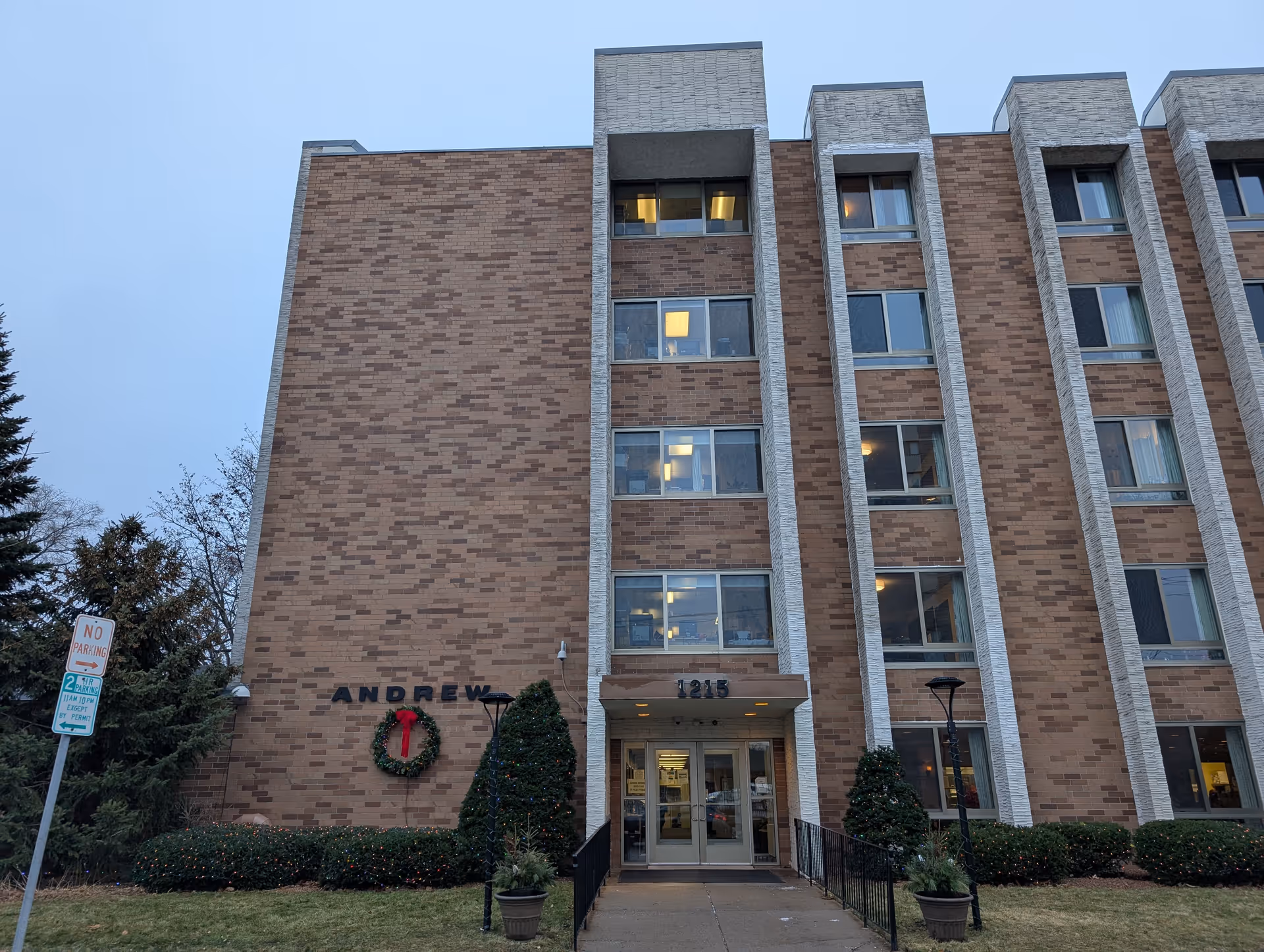 Exterior front view of a multi-story brick building named Andrew Residence with the address number 1215 above the entrance. The building has several windows, some with lights on inside. There is a wreath with a red bow on the wall near the entrance, and small decorated bushes and trees are visible in front of the building. A no parking sign is also visible on the left side of the image.